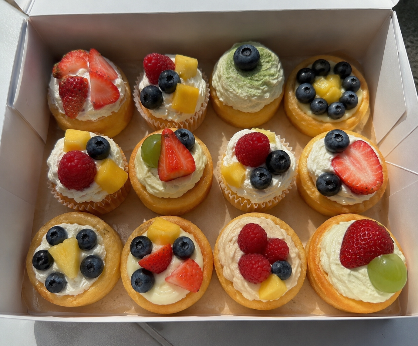A box of assorted fruit-topped cupcakes and tarts